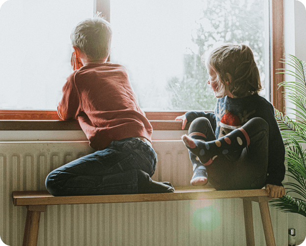 Two children sit near a radiator and look out the window at a dreary day