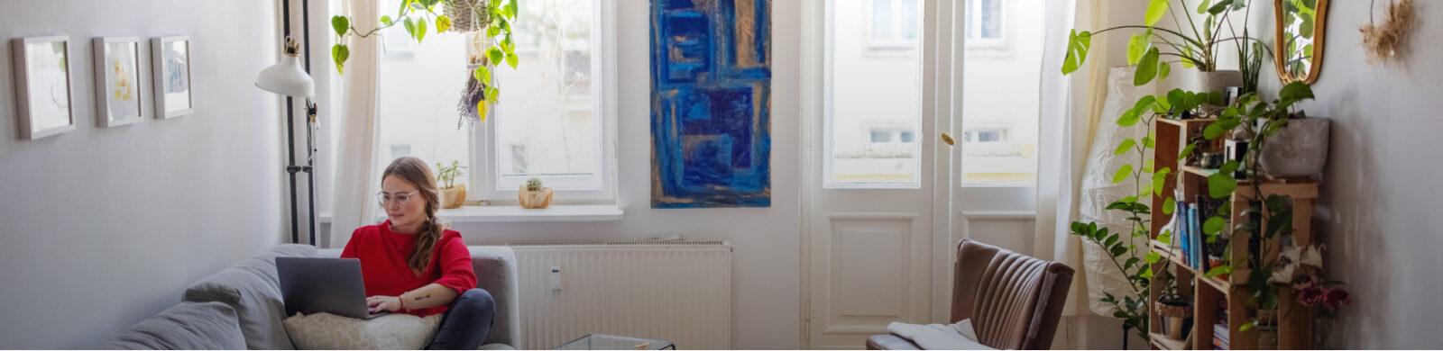 A woman in a red sweater sits in her living room on her couch as she works on her laptop.