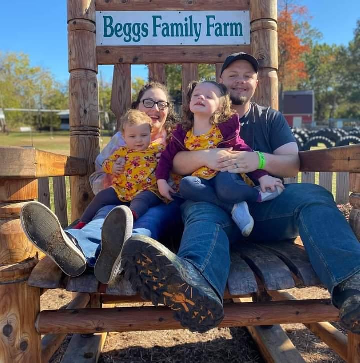 A Caucasian family of a man, woman and 2 young girls is seated outside in a large wooden rocking chair.
